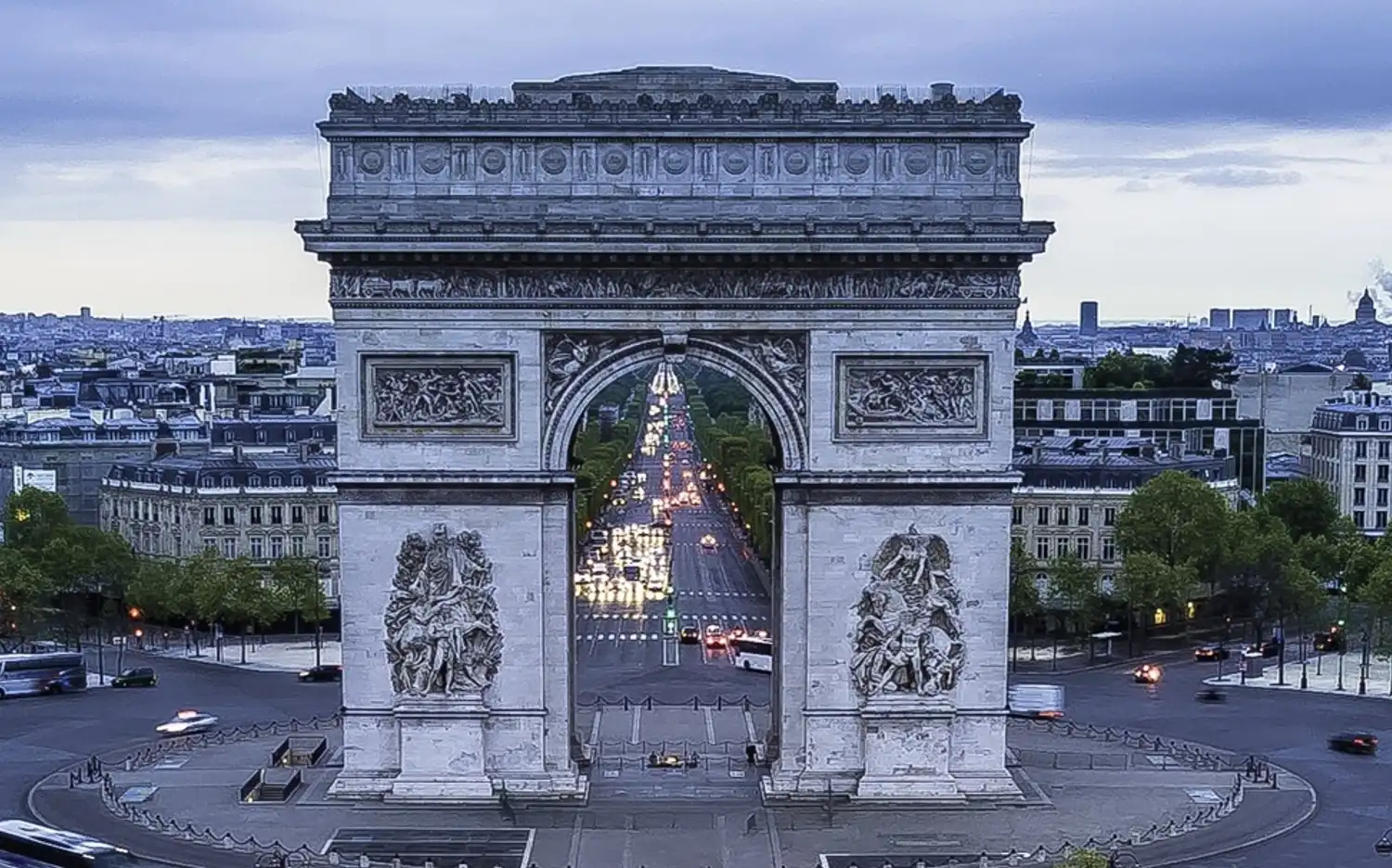 De Arc de Triomphe te midden van Parijse straten en gebouwen onder een bewolkte, grijze lucht.