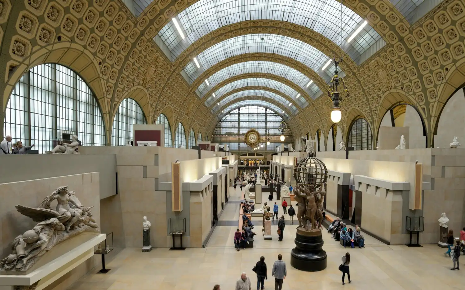A spacious museum hall with a vaulted glass ceiling, sculptures, and visitors walking and seated on benches.