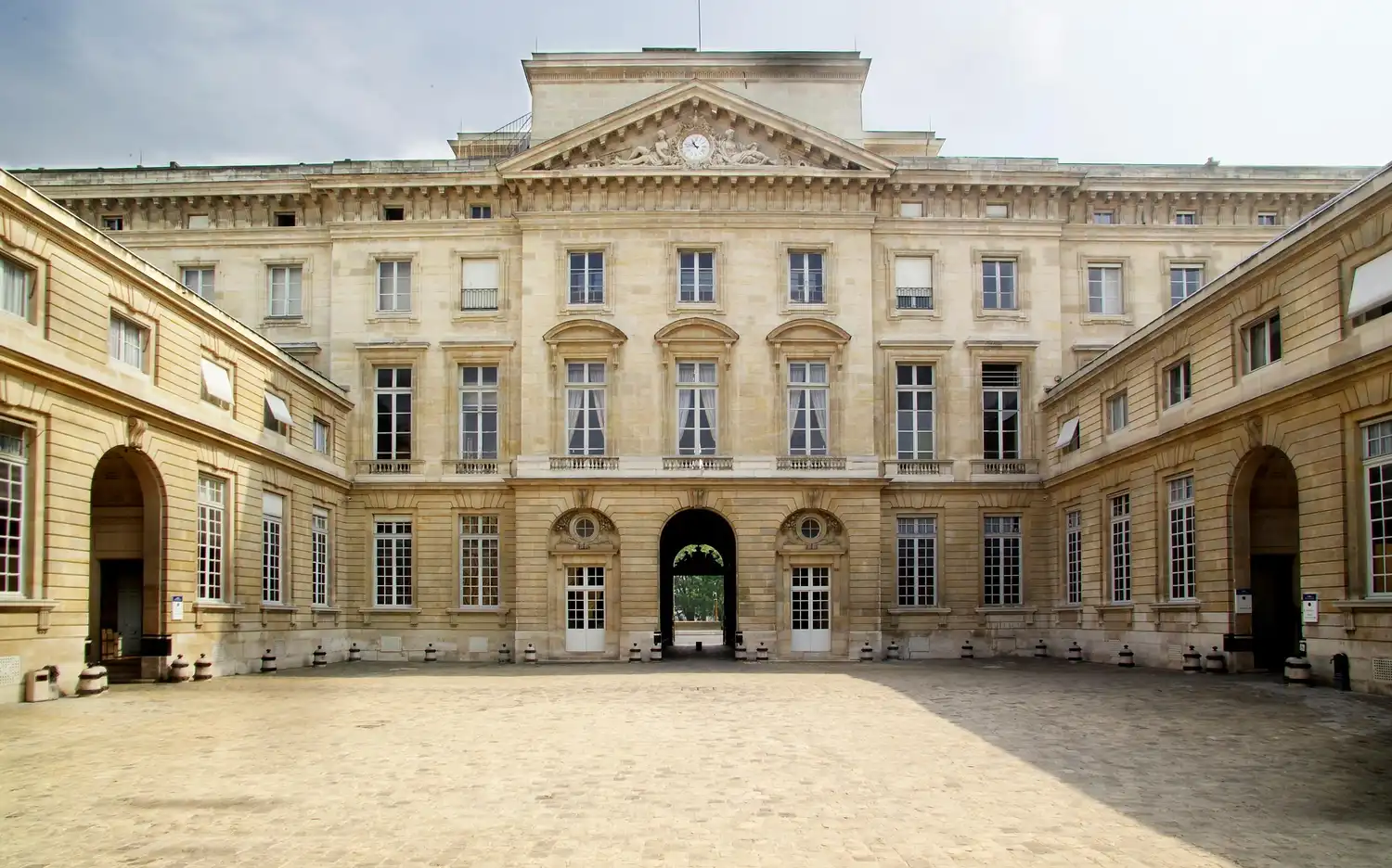 Groot, symmetrisch stenen gebouw van Monnaie de Paris met boogvensters en een centrale poort naar een binnenplaats.