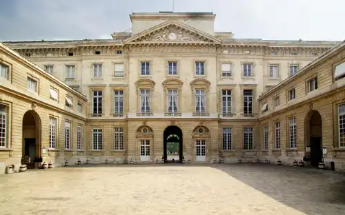 Neoklasycystyczna fasada Monnaie de Paris Monnaie de Paris: okazały, symetryczny kamienny budynek z łukowatymi oknami i wejściem na dziedziniec.