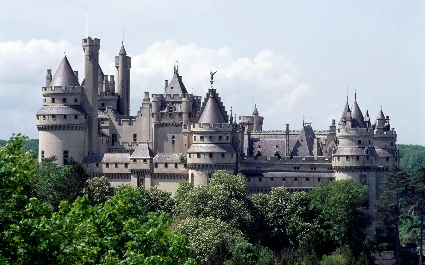 Zamek Château de Pierrefonds z licznymi wieżami i iglicami, otoczony zielenią na tle zachmurzonego nieba.
