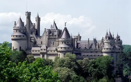 Gedetailleerd Château de Pierrefonds met torens en spitsen, omringd door groene bomen onder een bewolkte hemel.