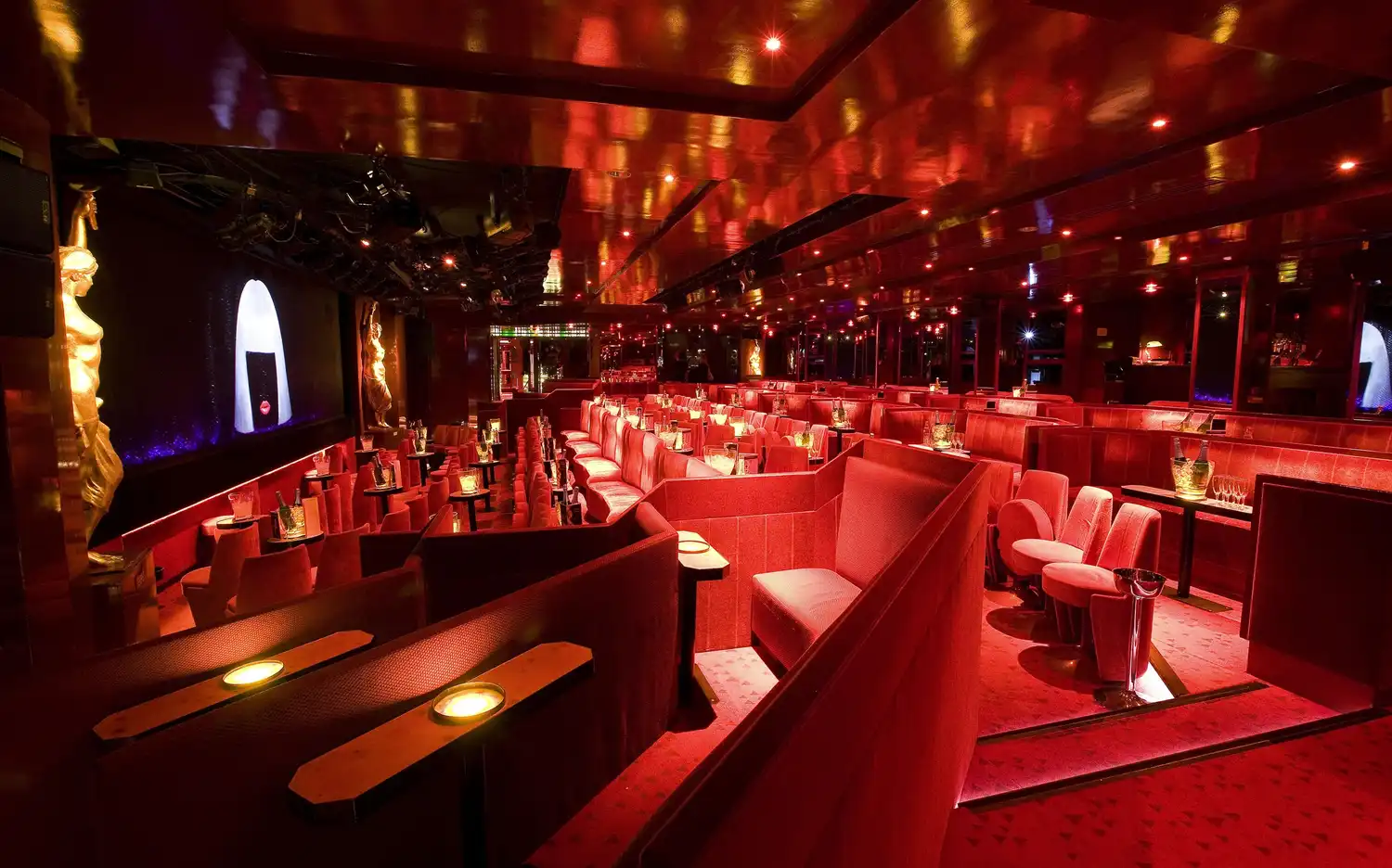 Salon faiblement éclairé avec des sièges rembourrés rouges, des tables individuelles, de petites lampes et des statues dorées, créant une atmosphère intime.