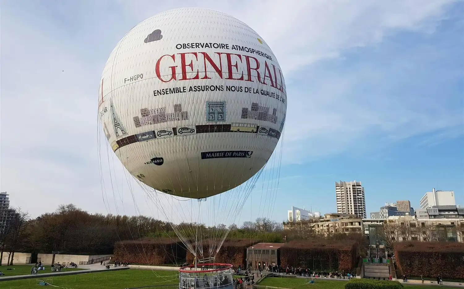Большой привязанный воздушный шар с рекламным текстом и дизайном городского пейзажа, расположенный в парке с людьми и зданиями на заднем плане. Большой привязанный воздушный шар с рекламным текстом и дизайном городского пейзажа, расположенный в парке с людьми и зданиями на заднем плане.