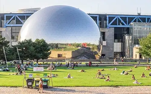 Kino Geode w Cité des sciences et de l'industrie Ludzie odpoczywają w słonecznym parku z drzewami i dużą metalową kopułą przy Cité des sciences et de l'industrie.