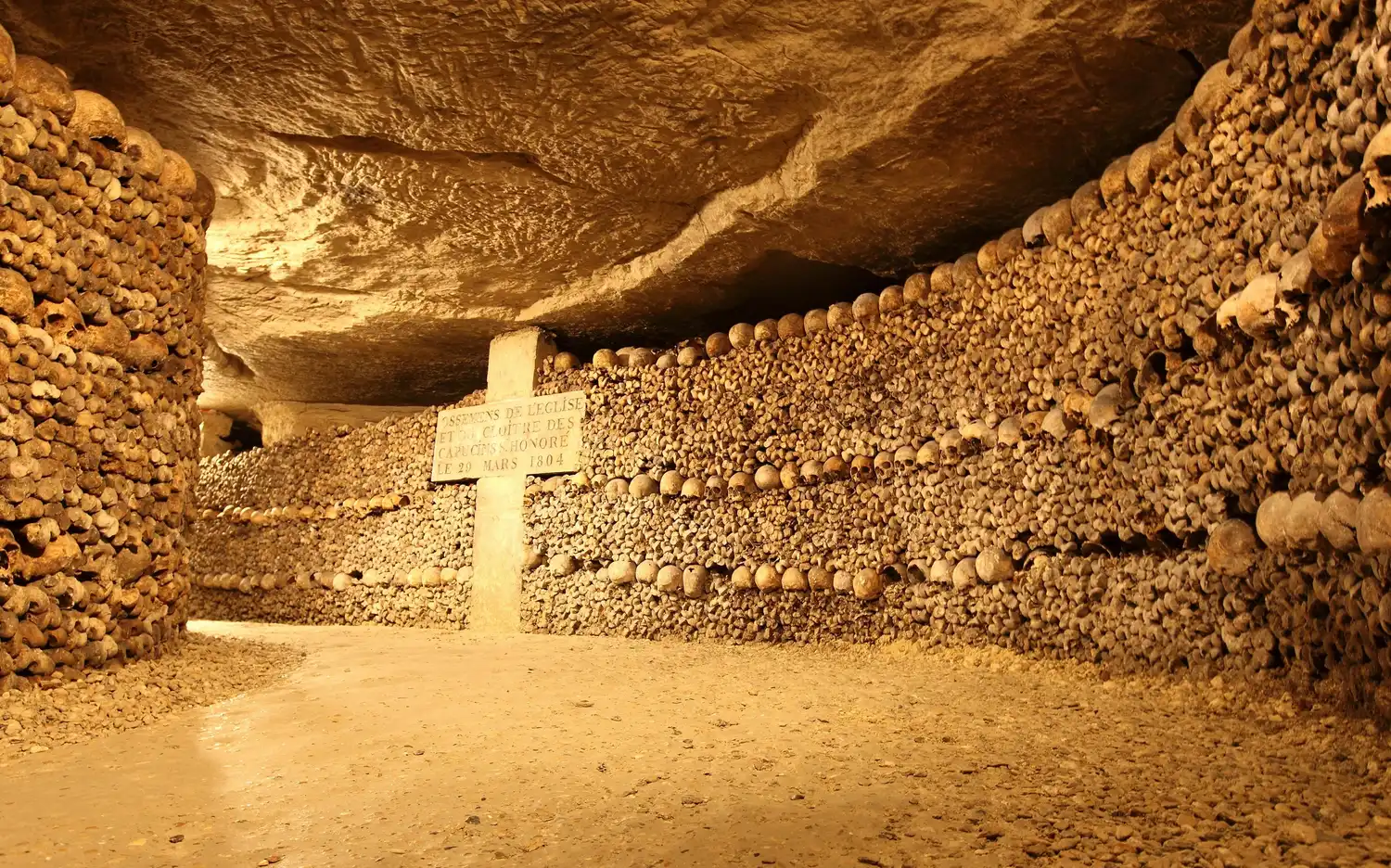 Un tunnel dont les murs sont constitués d'os et de crânes humains empilés, avec un pilier de pierre portant une inscription. Un tunnel dont les murs sont constitués d'os et de crânes humains empilés, avec un pilier de pierre portant une inscription.