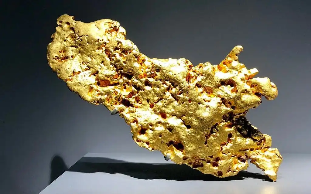 Grote, onregelmatige goudklomp met een ruwe textuur in de Geologie en Mineralogie Galerij op een grijze achtergrond.