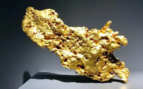 Grote, onregelmatige goudklomp met een ruwe textuur in de Geologie en Mineralogie Galerij op een grijze achtergrond.