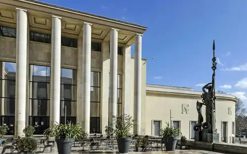 Extérieur d'un bâtiment néoclassique avec de hautes colonnes, de grandes fenêtres, des sièges extérieurs, des plantes en pot et une statue tenant une lance.