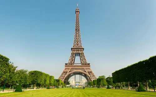 De Eiffeltoren gezien vanuit een grasveld met aan beide kanten gesnoeide bomen onder een helderblauwe lucht.