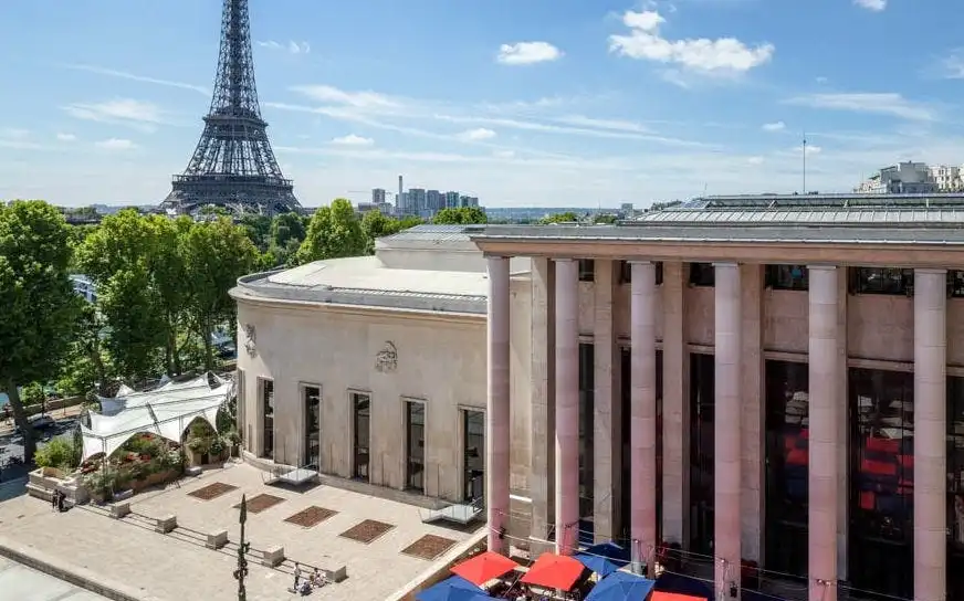 Monumentalne kolumny Le Palais de Tokio górują nad słonecznym dziedzińcem wypełnionym czerwonymi i niebieskimi parasolami, a w tle widoczna jest Wieża Eiffla.