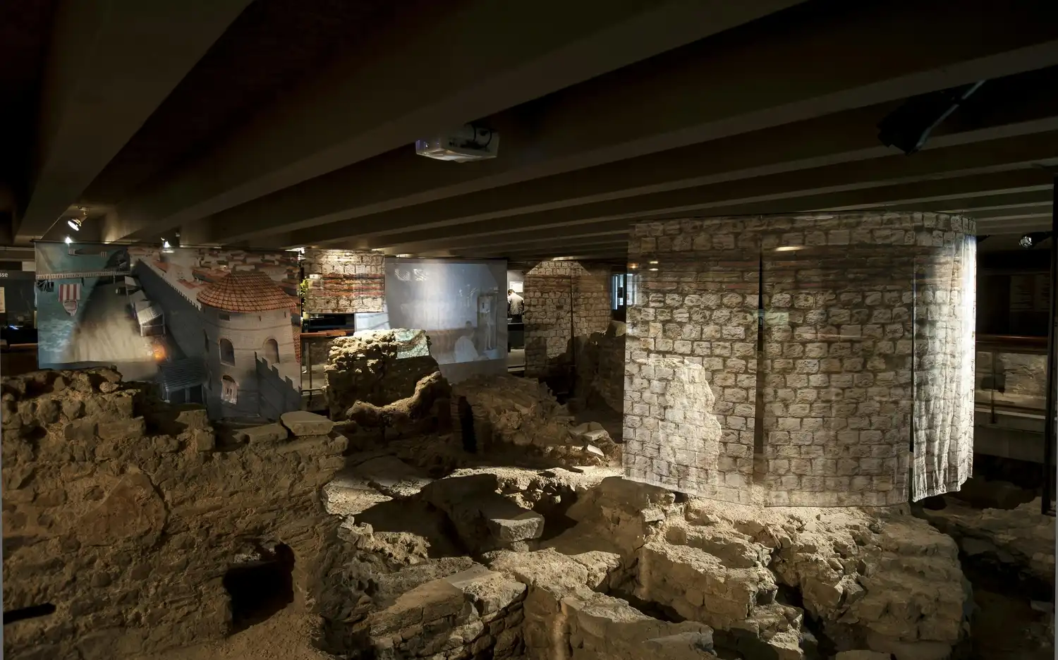 Des ruines anciennes sont exposées à l'intérieur avec des structures en brique et en pierre, et des images historiques sont projetées sur les murs.