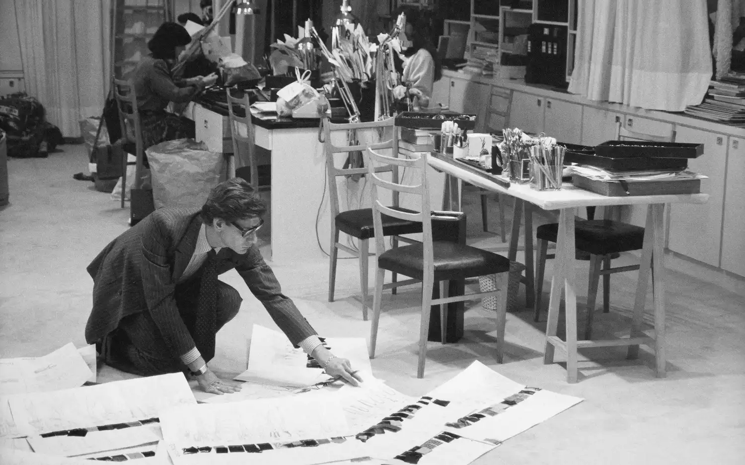 Un homme en costume examine de grands papiers sur le sol, tandis que deux femmes travaillent à des bureaux dans une pièce encombrée.