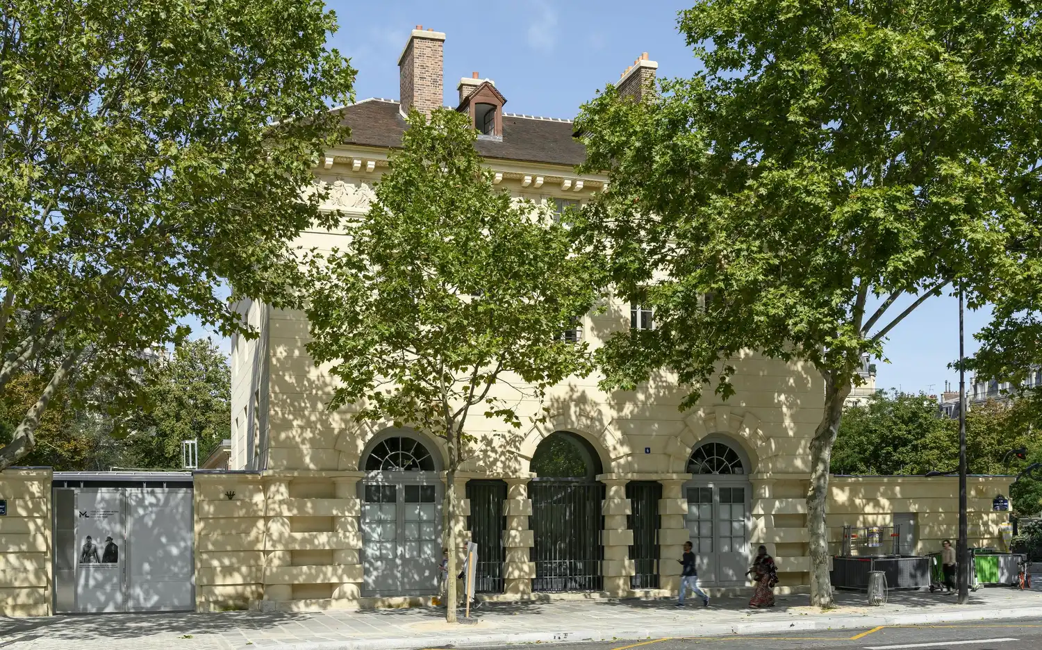 Bâtiment en pierre avec arcades et fenêtres, partiellement masqué par des arbres feuillus. Deux piétons marchent sur le trottoir. Bâtiment en pierre avec arcades et fenêtres, partiellement masqué par des arbres feuillus. Deux piétons marchent sur le trottoir.