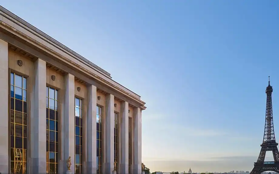 Un bâtiment avec des colonnes sur la gauche, un trottoir à motifs et la Tour Eiffel en arrière-plan dans un ciel bleu clair.