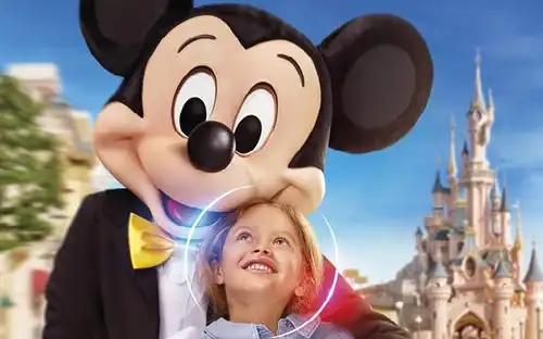 Un enfant sourit à une personne déguisée en Mickey Mouse, avec un château de conte de fées et des bâtiments colorés en arrière-plan.