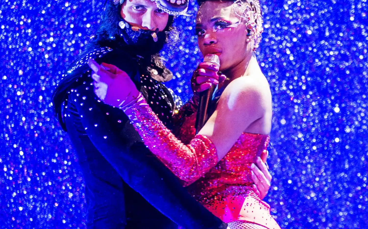 Sur une scène bleue pailletée, un artiste portant un masque de cheval embrasse une chanteuse vêtue d'une tenue rouge étincelante. Sur une scène bleue pailletée, un artiste portant un masque de cheval embrasse une chanteuse vêtue d'une tenue rouge étincelante.