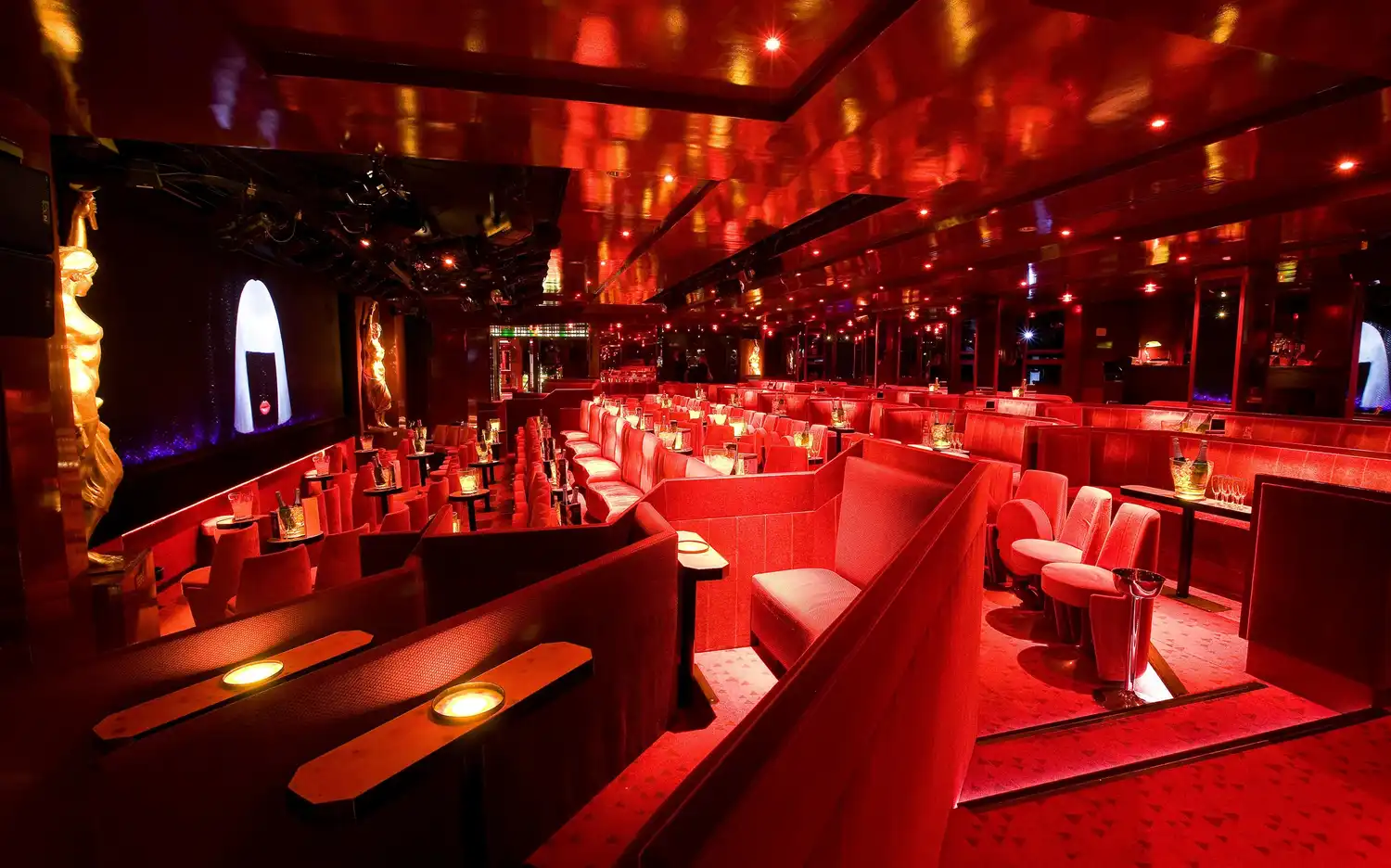 Gedimd verlichte lounge met rode bekleding, tafeltjes, lampjes en gouden beelden bij Crazy Horse Parijs.