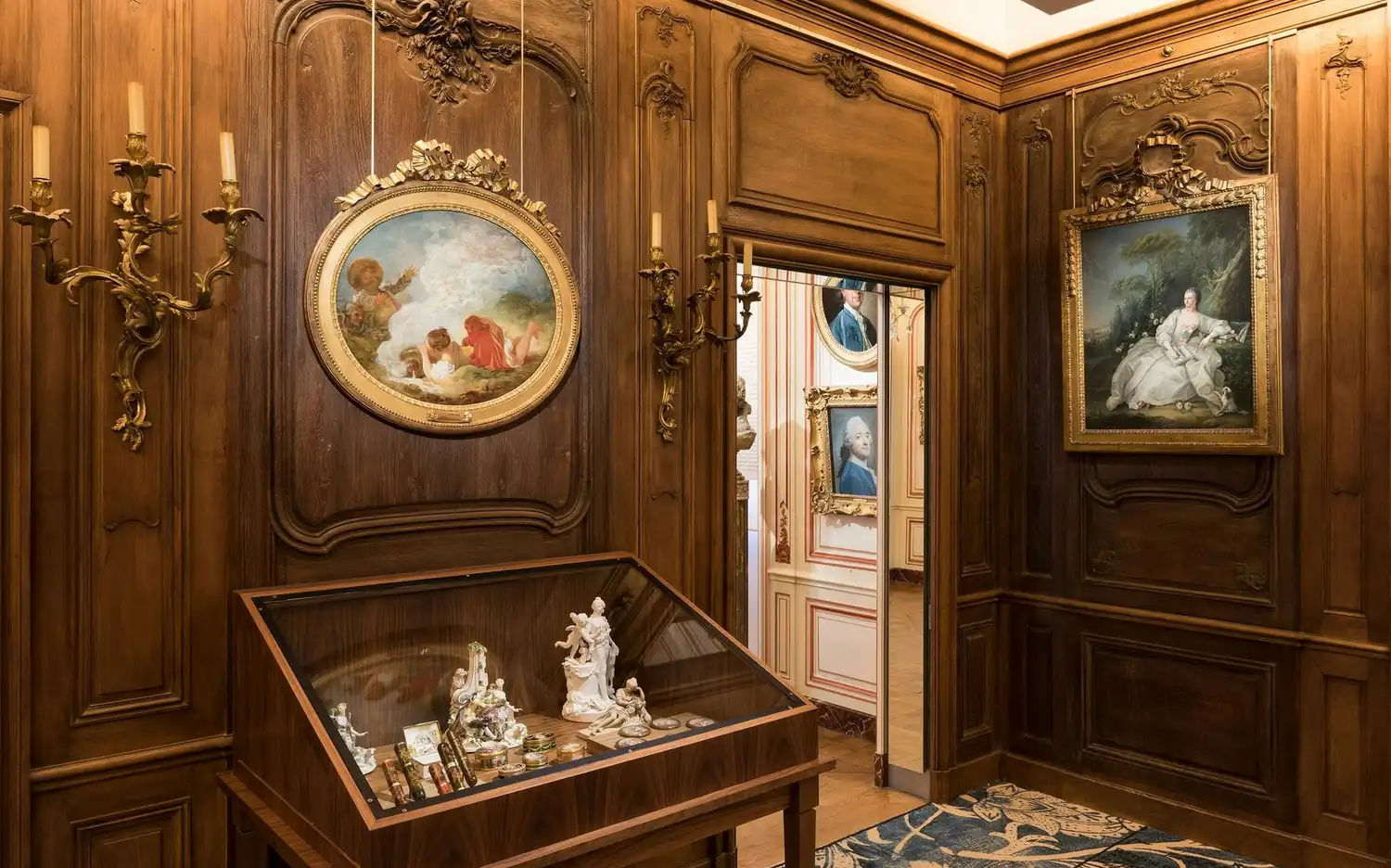 Pièce ornée de boiseries, d'appliques dorées, de deux tableaux encadrés et d'une vitrine contenant des figurines en porcelaine et de petits objets.