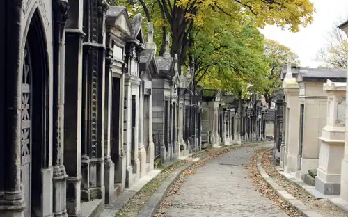 Kasseienpad op Begraafplaats Père Lachaise omzoomd door mausoleums en bomen met groene en gele bladeren.