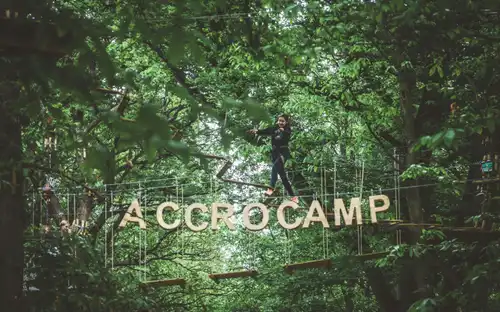 Een persoon balanceert op een hoog touwenparcours bij AccroCamp: Paris-Jablines boven grote houten letters.