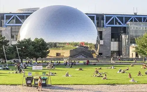 Des personnes se détendent dans un parc herbeux avec des arbres et une grande structure métallique en forme de dôme en arrière-plan par une journée ensoleillée.