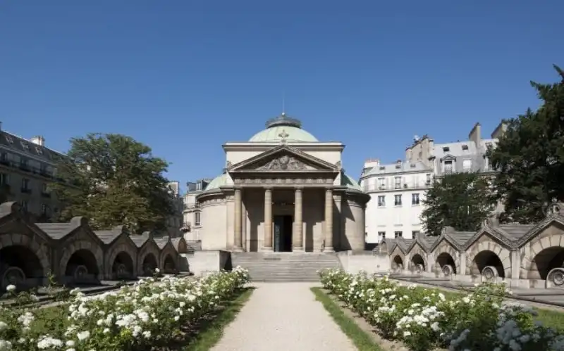 Un bâtiment classique avec des colonnes et un dôme, entouré de fleurs et d'arbustes blancs, sur fond de ciel bleu clair.