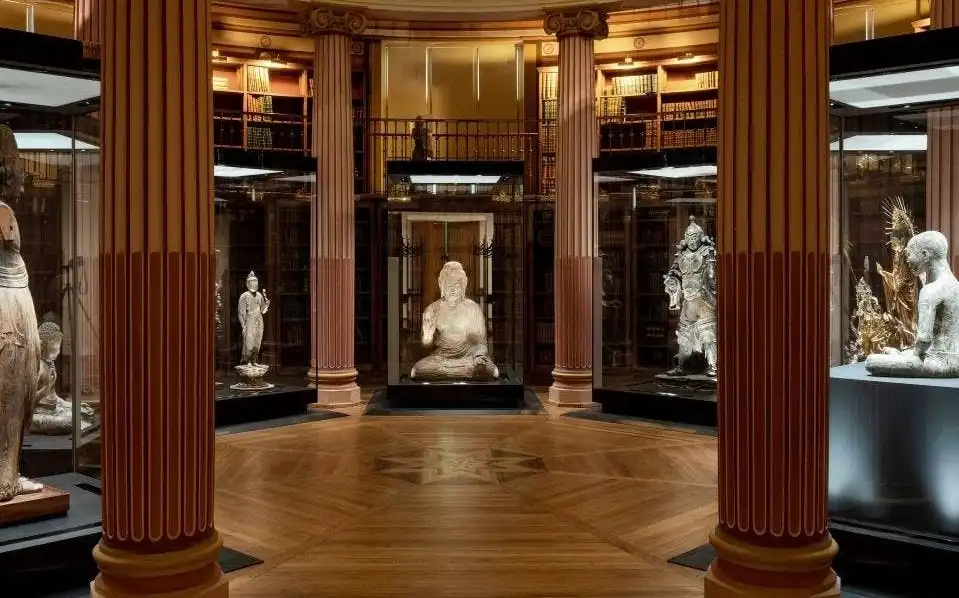 Trzy rzeźby w gablotach w sali Musée National des Arts Asiatiques Guimet, otoczone regałami i zdobionymi kolumnami.
