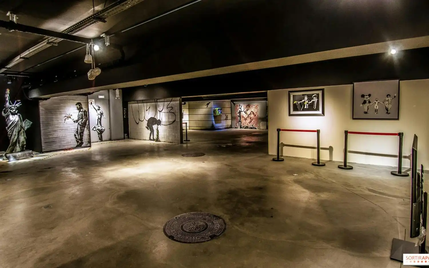 Une galerie faiblement éclairée présentant de l'art de rue, notamment des peintures murales, des gravures encadrées et une statue, avec des barrières délimitant certaines zones. Une galerie faiblement éclairée présentant de l'art de rue, notamment des peintures murales, des gravures encadrées et une statue, avec des barrières délimitant certaines zones.