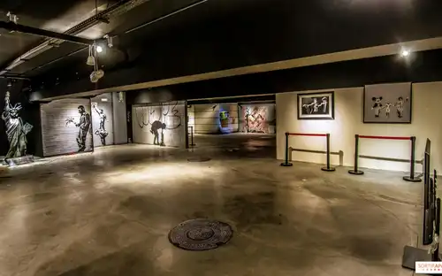 Une galerie faiblement éclairée présentant de l'art de rue, notamment des peintures murales, des gravures encadrées et une statue, avec des barrières délimitant certaines zones.