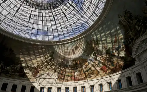 Un grand dôme de verre avec une structure métallique complexe, qui projette des ombres sur une peinture murale représentant des scènes historiques et mythiques.