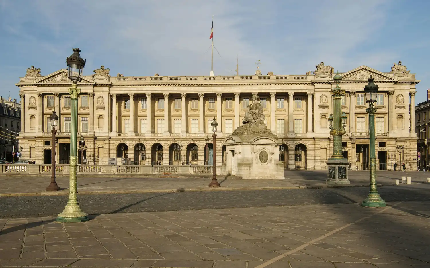 Историческое здание с французским флагом, богато украшенным фасадом, колоннами, зелеными фонарными столбами и статуей на открытой площади с каменным покрытием.
