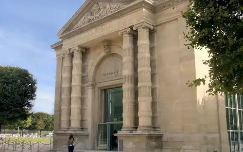 Un bâtiment en pierre orné de grandes colonnes et portant l'inscription "Orangerie" au-dessus de l'entrée. Une personne se tient près de la porte.
