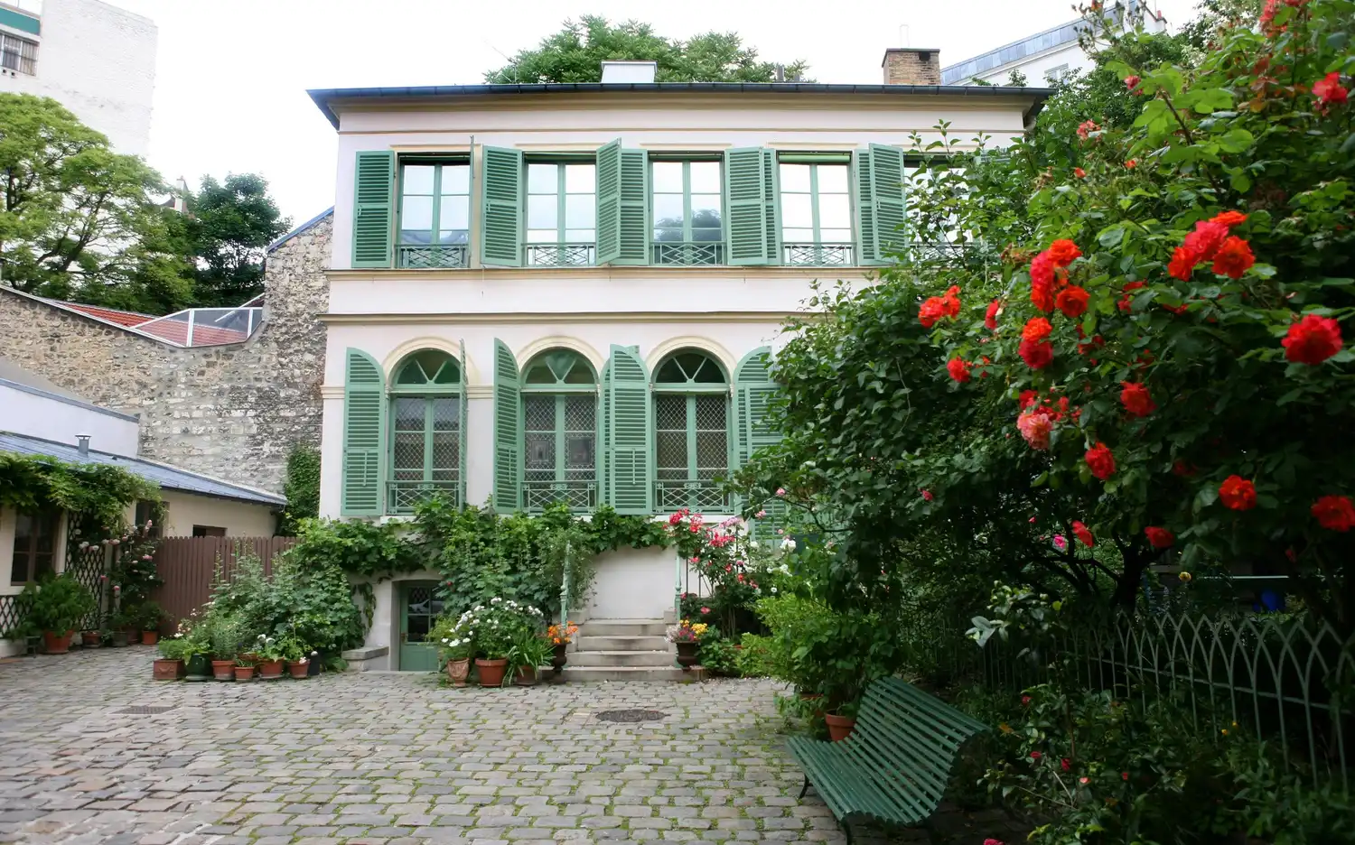 Brukowny dziedziniec Musée de la Vie Romantique z zieloną ławką, czerwonymi kwiatami w donicach i zielonymi okiennicami.
