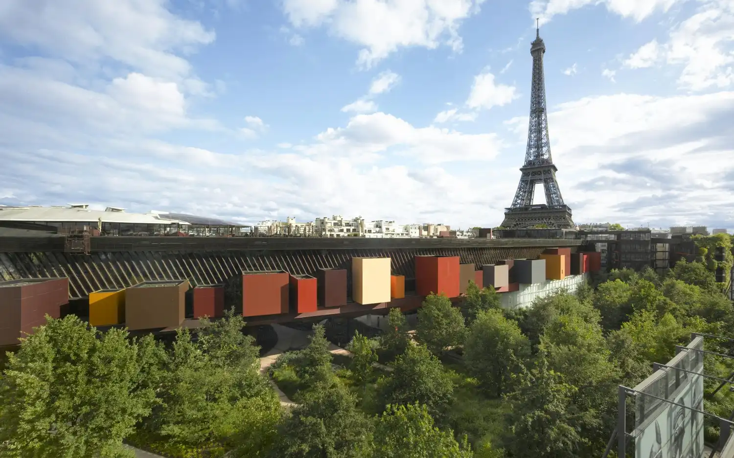 Bâtiments colorés en forme de blocs entourés d'arbres verdoyants, avec la Tour Eiffel visible à l'arrière-plan sous un ciel partiellement nuageux.