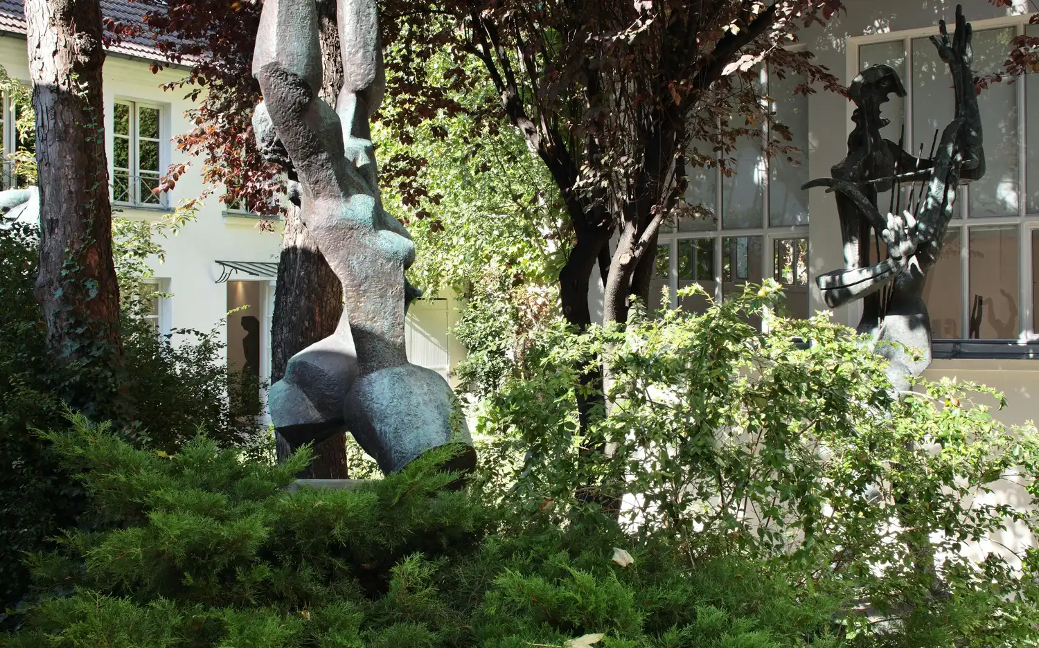 Бронзовые статуи абстрактных человеческих фигур в зеленом саду со зданиями и окнами на заднем плане.