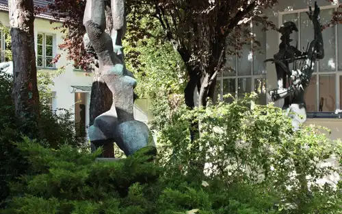 Statues en bronze de figures humaines abstraites dans un jardin verdoyant, avec des bâtiments et des fenêtres en arrière-plan.