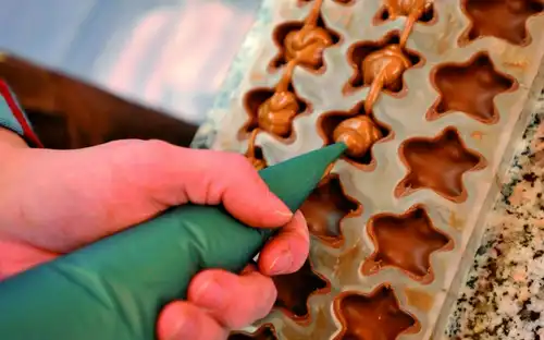 Une main remplissant de chocolat des moules en forme d'étoile à l'aide d'une poche à douille verte sur un comptoir en granit.