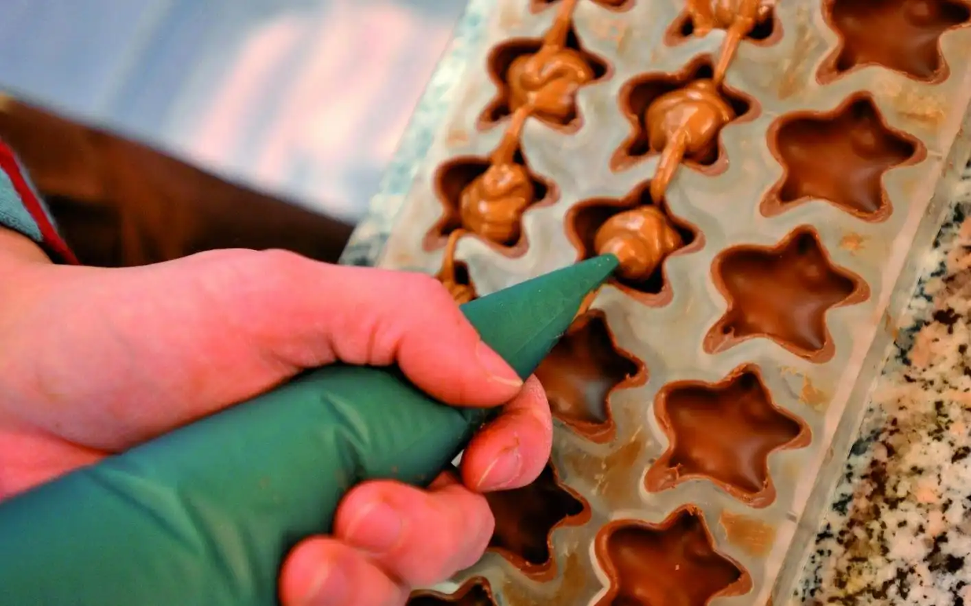 Une main remplissant de chocolat des moules en forme d'étoile à l'aide d'une poche à douille verte sur un comptoir en granit.