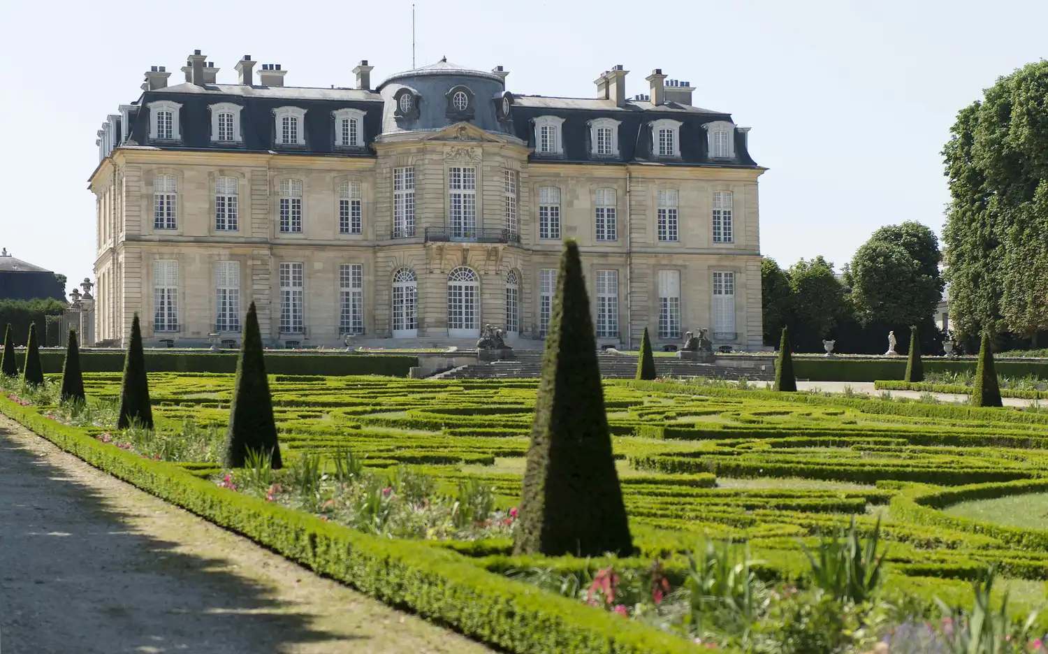 Zabytkowy gmach Château de Champs-sur-Marne z zadbanymi ogrodami, przyciętymi żywopłotami i stożkowatymi krzewami.