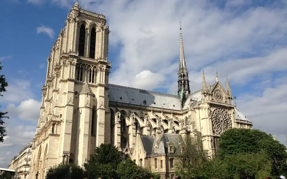 Tłum przed Katedrą Notre-Dame w Paryżu pod częściowo zachmurzonym niebem z widocznymi wieżami i iglicą.