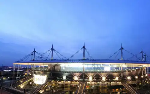 Stade illuminé au crépuscule avec des projecteurs visibles, plusieurs rampes d'accès et un ciel bleu crépusculaire.