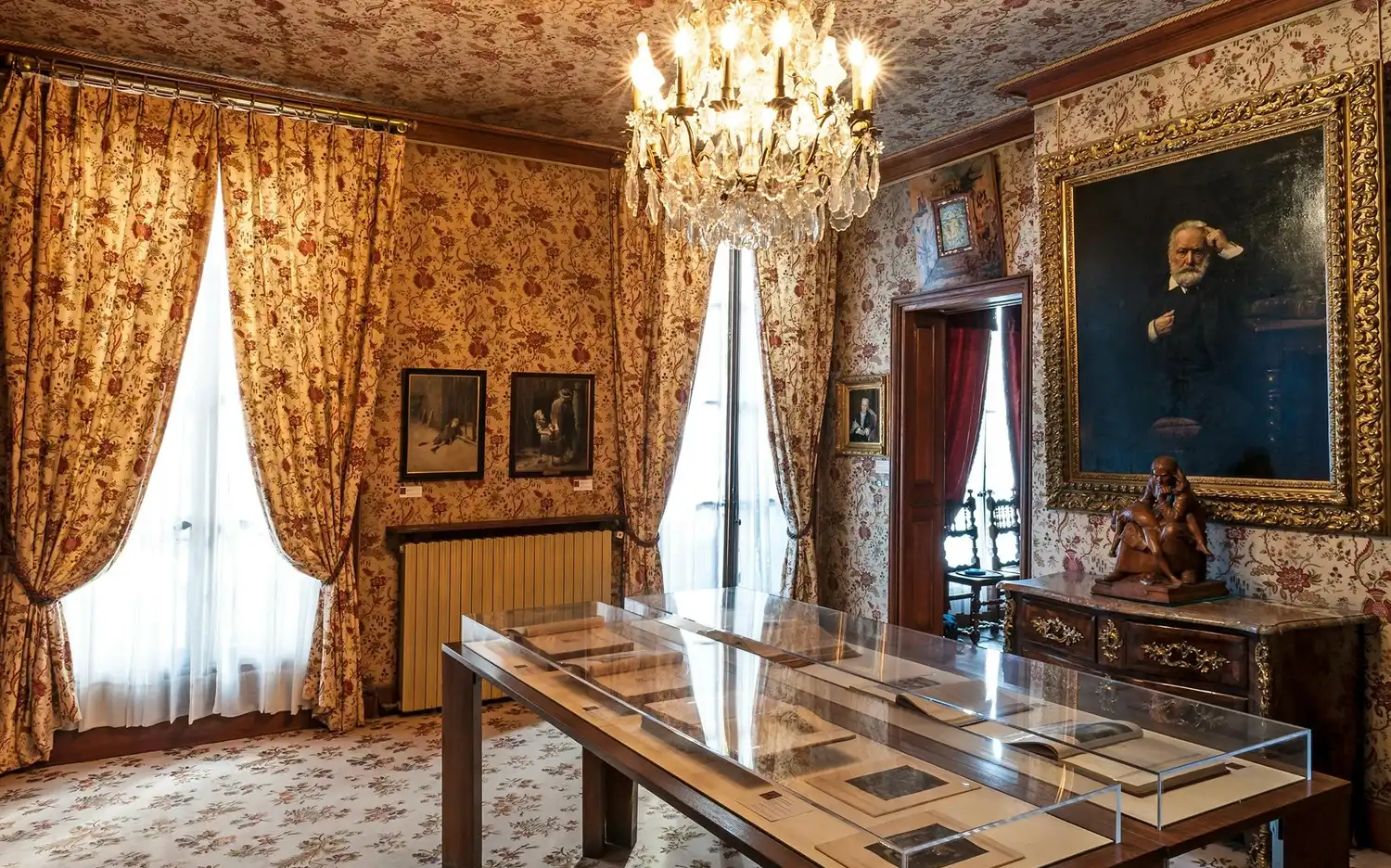 Pièce ornée d'un papier peint floral, d'un lustre, de portraits encadrés, de meubles anciens et d'une table d'exposition avec des documents. Pièce ornée d'un papier peint floral, d'un lustre, de portraits encadrés, de meubles anciens et d'une table d'exposition avec des documents.