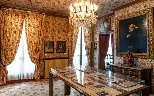 Pièce ornée d'un papier peint floral, d'un lustre, de portraits encadrés, de meubles anciens et d'une table d'exposition avec des documents.