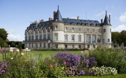 Château de Rambouillet met grijze daken en ronde torens, omringd door kleurrijke bloementuinen onder een blauwe lucht.