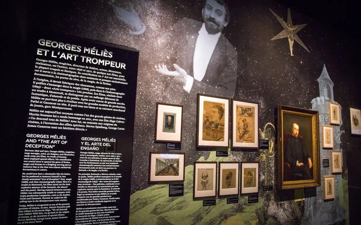 Exhibit wall with text about Georges Méliès in French, English, and Spanish, surrounded by various framed portraits and illustrations. Exhibit wall with text about Georges Méliès in French, English, and Spanish, surrounded by various framed portraits and illustrations.
