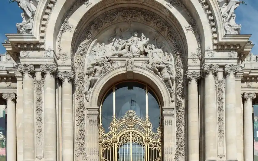 Une grande entrée voûtée avec des sculptures complexes, de grandes portes dorées, flanquées de sculptures et de colonnes sous un ciel bleu.