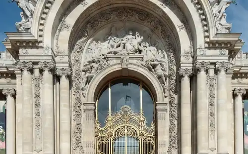 Une grande entrée voûtée avec des sculptures complexes, de grandes portes dorées, flanquées de sculptures et de colonnes sous un ciel bleu.