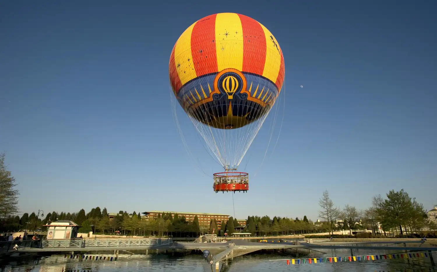 A colorful hot air balloon is tethered above a pond with a dock, surrounded by trees and buildings under a clear blue sky.