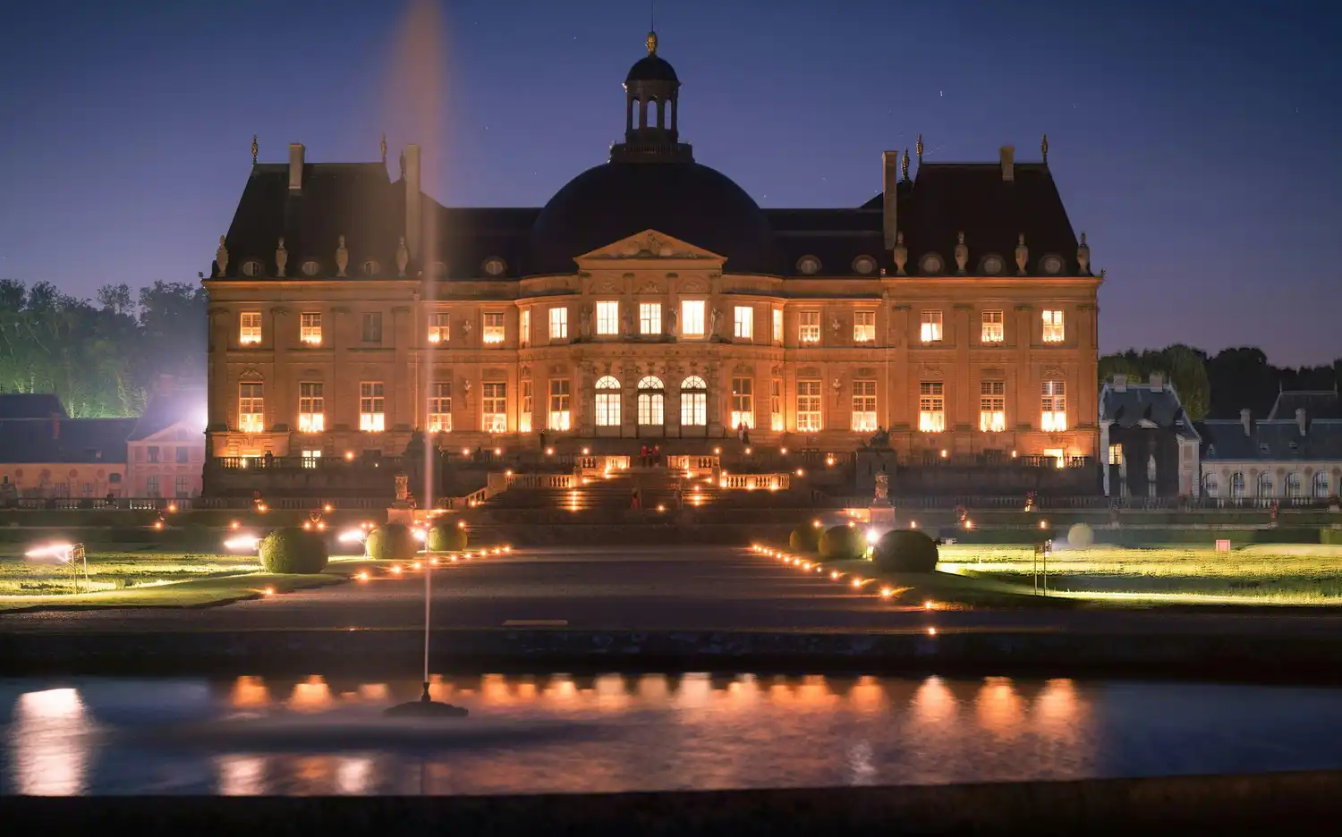 Verlicht Kasteel van Vaux-le-Vicomte bij nacht met tuinen, fontein en waterreflectie op de voorgrond.
